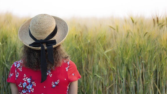 Rückansicht einer Frau, die einen Strohhut und ein rotes Sommerkleid trägt und vor einem Feld steht.
