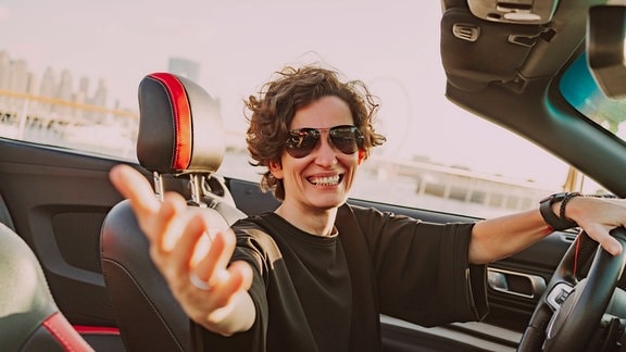 Glückliche Frau mit Sonnenbrille gestikuliert an einem sonnigen Tag im Cabrio.