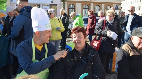 Ein Mann mit Kochmütze hält einer Frau ein Mikrofon vor en Mund.