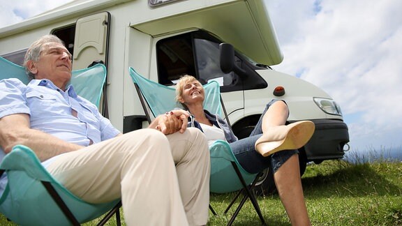 Eine Frau und ein Mann sitzen in Campingstühlen vor einem Wohnmobil.