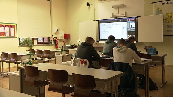 Vier Schüler sitzen in einem Klassenraum und folgen dem Unterricht via Videoübertragung auf einer digitalen Tafel.