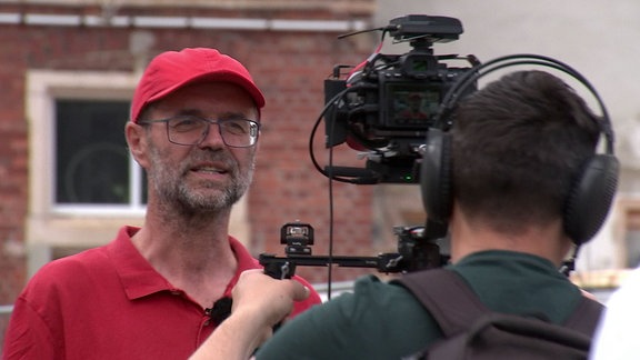 Mann mit rotem Hemd und roter Mütze lässt sich von Kamera filmen.