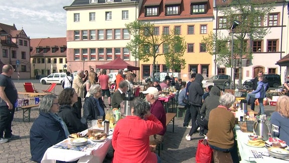 Menschen sitzen an mehreren Tischen auf einem Marktplatz.