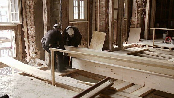 Zwei Personen auf einer Baustelle heben einen großen Holzbalken.