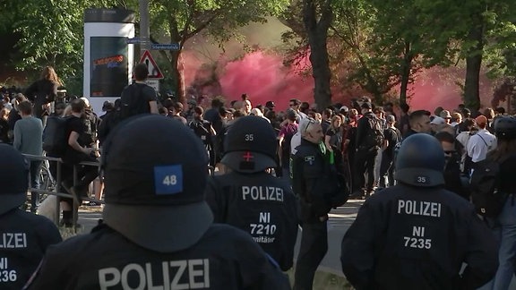 Städtische Szene mit Polizei im Vordergrund und dichten, roten Rauchschwaden im Hintergrund