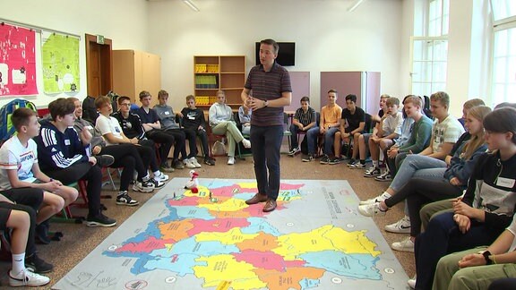 Ein Lehrer steht auf einer großen, bunten Landkarte, umringt von Schüler*innen.