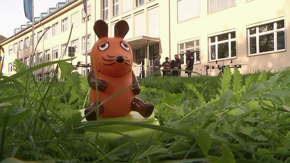 Eine Figur der Sendung mit der Maus sitzt im Gras.