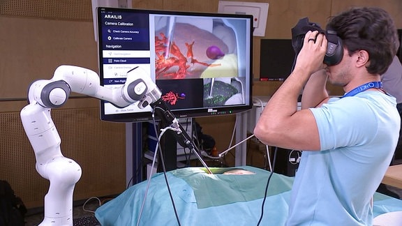 Ein Mann mit VR Brille schaut auf einen Monitor, ein Roboterarm daneben während einer simulierten OP