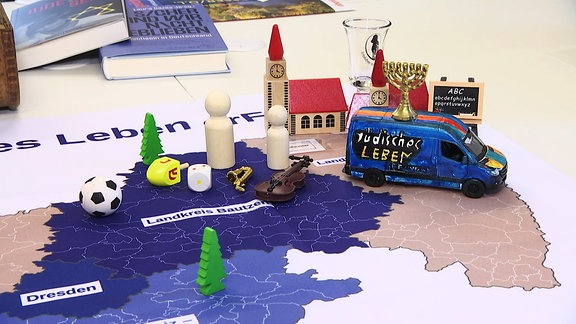 Ein Spielzeugbus auf dem Jüdisches Leben steht steht neben weiteren Spielfiguren auf einer Landkarte.