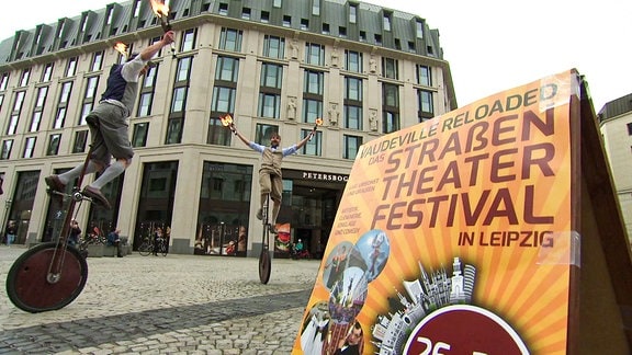 Jongleure auf Einrädern, davor ein Schild zu einem Straßenfestival