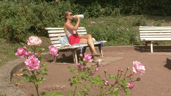 Eine Frau trinkt auf einer Parkbank Wasser
