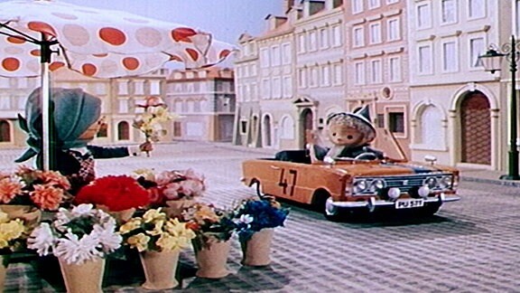 Der Sandmann fährt mit einem Auto an einem Blumenstand vorbei.