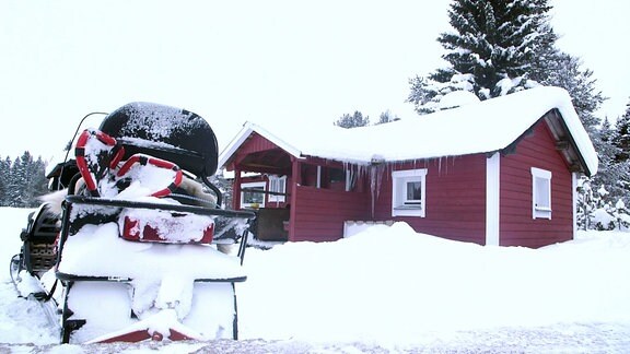 Verschneite Hütte, davor ein Motorschlitten mit Anhänger.