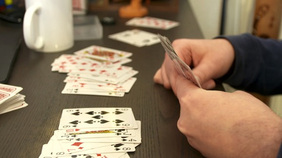 Spielkarten liegen auf einem Tisch, weitere Karten werden von zwei Händen gehalten