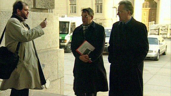 Ein Mann mit Bart und heller Jacke steht vor einem Gebäude und spricht zwei Männer links von der Seite an. Einer der beiden trägt Akten unter dem Arm. 