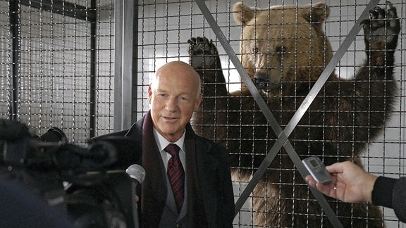 Vor dem Bärenkäfig ist "der Bär" los. Vor laufenden Kameras erklärt Staatssekretär Bredow (Dietrich Hollinderbäumer) der Presse stolz, dass es nur seinem Einsatz zu verdanken ist, dass der Braunbär nicht getötet, sondern gefangen und gesundgepflegt wurde. Er kann wieder ausgewildert werden.