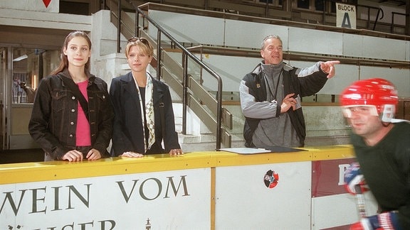 Lisa (Marie-Ernestine Worch, li.) und Tanja (Tanja Szewczenko) beobachten das Training der Pinguins. Trainer Sänger (Jürgen Heinrich) gibt Anweisungen auf die Eisfläche.