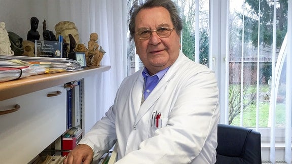 Prof. Werner Schunk hat ein Arztkittel um und schaut, neben einem Aktenschrank stehend, in die Kamera.