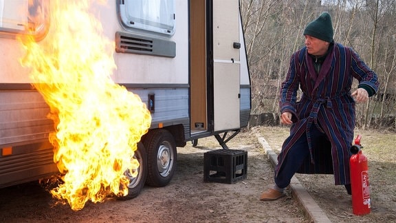 Klaues (Henry Hübchen) Wohnwagen steht in Flammen. Klaue stürzt raus, greift zum Feuerlöscher und versucht, zu löschen.