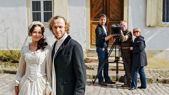 Minna Wagner (Ivonne Braasch) und Richard Wagner (Norman Stoffregen) lächeln altmodisch bekleidet nach vorn. Im Hintergrund steht ein Kamerateam. 