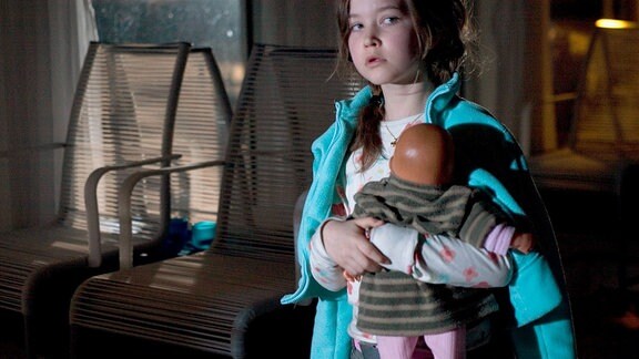 Die kleine Cleo (Nadja Qrunell), welche den Mord an ihrer Mutter beobachtet hat, steht mit einer Puppe auf dem Arm da.