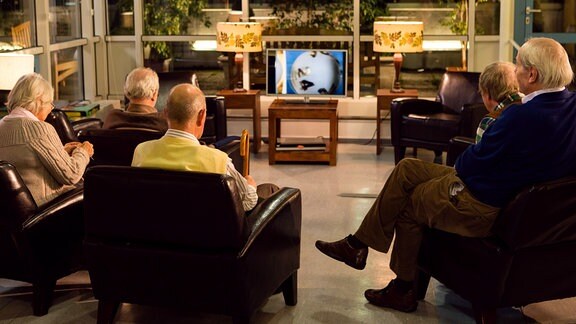 Sechs ältere Menschen sitzen gemeinsam in einem Raum und sehen fern. 
