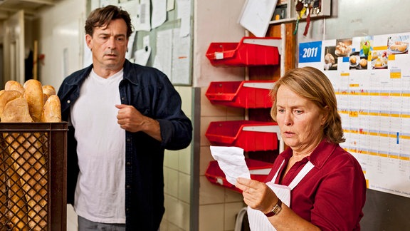 Oliver Maibach (Thomas Sarbacher) und seine Mutter Elisabeth (Petra Kelling)stehen in einer Bäckerei. Die Mutter hält ein Papier in der linken Hand und starrt darauf.