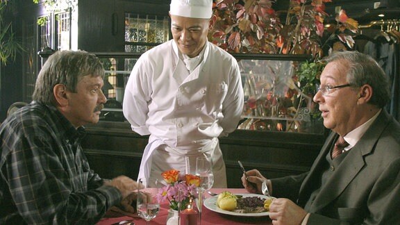 Schmücke (Jaecki Schwarz) und Schneider (Wolfgang Winkler) am Tisch neben japanischem Koch.