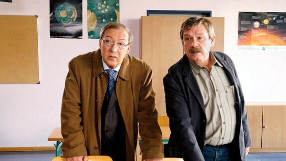Schmücke (Jaecki Schwarz, links) und Schneider (Wolfgang Winkler, rechts) stehen in einem Klassenzimmer hinter einem Schultisch.