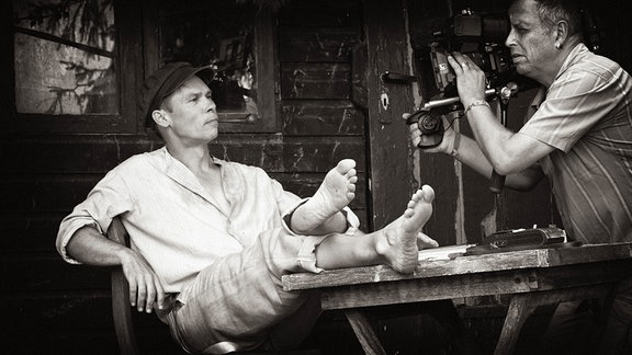 Ein Mann in einfacher Kleidung des frühen 20. Jahrhunders sitzt auf einem Stuhl, die Füße auf dem Tisch überschlagen. Ein Kameramann filmt ihn dabei.