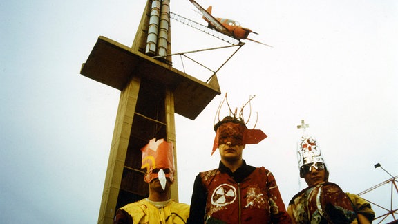 Drei Künstler des Kunstkollektivs AG Geige vor einem Fahrgeschäft. Die Künstler tragen Masken und bunte Kostüme.