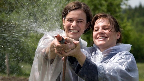 Ellen (Maria Simon) und ihre Schwester Leonie (Annika Kuhl, l.) halten  gemeinsam einen Wasserschlauch.