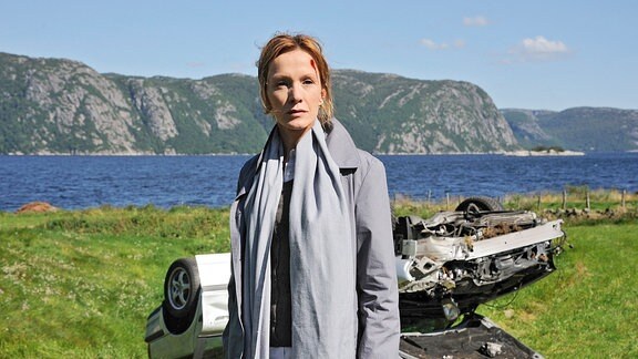 Brigitta (Katja Flint). Hinter ihr ein beschädigtes Auto, das auf dem Dach liegt.