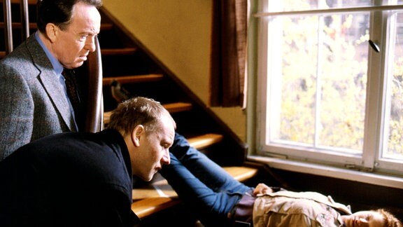 Die Hauptkommissare Ehrlicher (Peter Sodann, links) und Kain (Bernd Michael Lade) blicken an  Tatort auf Rotkäppchen (Laura Laß), das am Fu0e einer Treppe liegt..