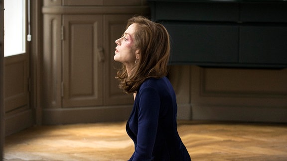 Michèle Leblanc (Isabelle Huppert) sitzt auf einem Parkett-Fußboden, Neben ih zerbrochene Gläser.