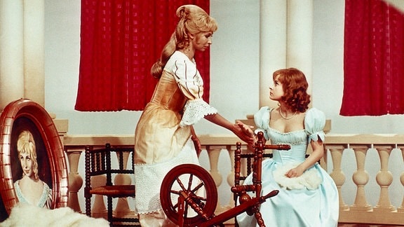 Szene aus einem Film, in dem ein Mädchen in einem hellblauen Kleid hinter einem Spinnrad sitzt, neben ihm steht eine Frau in einem weißen Kleid mit goldenem Besatz und hält ihm die Hand