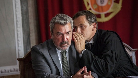 Tiago Zarco (André Gago)wird von seinem Anwalt Abel Ferreira (Ben Bela Böhm) etwas zugeflüstert; Zarco blickt mürrisch.