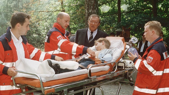 Kommissar Schmücke (Jaecki Schwarz) erkundigt sich bei Rettungssanitätern nach dem Zustand des kleinen Jonas Köhler (Frederik Abrahams, auf einer Krankentrage).