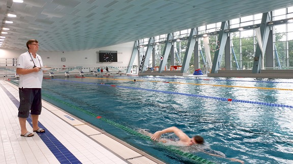 Schwimmtrainer Frank Embacher steht am Beckenrand einer Schwimmhalle. 