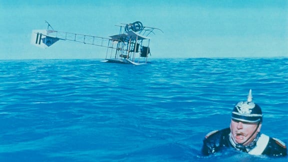 Oberst Manfred von Holstein (Gert Fröbe) in Uniform und schwimmend. Im Hintergrund treibt sein abgestürztes Flugzeug auf dem Wasser.