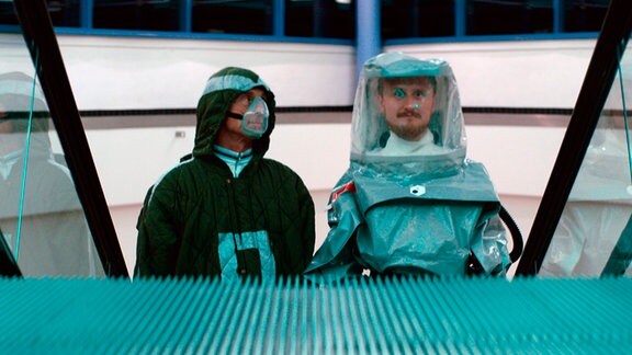 Falter (André M. Hennicke, li.) und Dave (Michael Kranz, re.) stehen in Schutzkleidung auf einer Rolltreppe. 