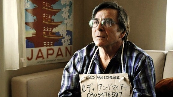 Damit Rudi (Elmar Wepper) in der Millionenstadt Tokio nicht verloren geht, wird ihm ein Schild mit Adresse um den Hals gehängt.