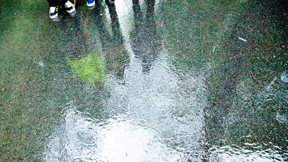 Regenfilm auf Asphalt. Am oberen Rand sind beschuhte Füße mehrer Personen zu entdecken.