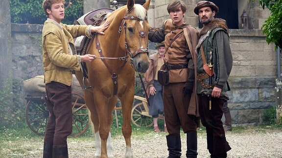 Prinz Lennard (Gustav Schmidt, M), Jäger Baldur (Luca Zamperoni, r) und der Stallbursche Gunther (Marcel Gewehr, l) - neben einem Pferd stehend, blicken an der Kamera vorbei.