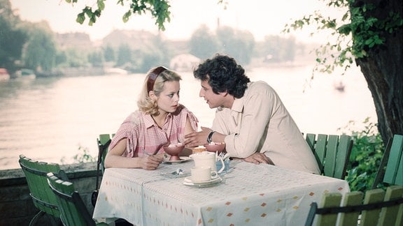 Szene aus der Serie "Jockei Monika": Monika Zeller (Miroslava Šafránková) und Udo Bujoldl (Hilmar Eichhorn) sitzen an einem Tisch unter einem Baum