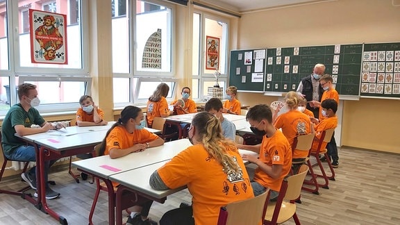 Eine Gruppe von elf Kindern - die meisten in orangen T-Shirts - sitzen in einem Klassenzimmer, ein älterer Mann steht an der Tafel.