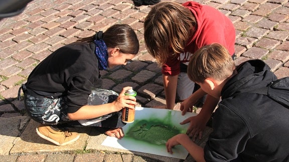 Kinder besprühen ein Blatt Papier mit Farbe auf der Straße.