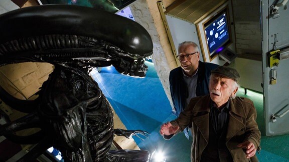 Zwei ältere Männer stehen vor einer "Alien"-Plastik.
