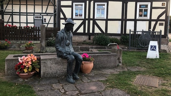 Eine Denkmal an einem Brunnen, auf dem eine Statur angebracht ist. Diese stellt eine sitzende Person mit Hut und Halskette mit Muschelanhänger dar. Im Hintergrund Fachwerkhäuser.