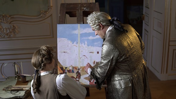 Ein Mann mit Kleidung aus dem 18. Jahrhundert zeigt einem anderen Mann in ähnlicher Kleidung etwas auf einem Bild, das auf einer Staffelei steht.
